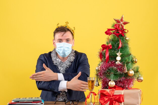 Hombre de negocios de vista frontal cruzando las manos sentado en la mesa cerca del árbol de Navidad y presenta sobre fondo amarillo