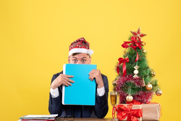 Hombre de negocios de vista frontal cubre su rostro con una carpeta de archivos sentado en la mesa cerca del árbol de Navidad y presenta sobre fondo amarillo