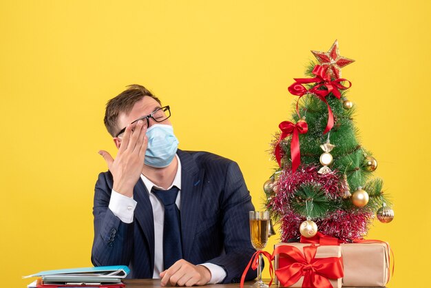 Hombre de negocios de vista frontal que cubre su ojo con la mano sentado en la mesa cerca del árbol de Navidad y presenta sobre fondo amarillo