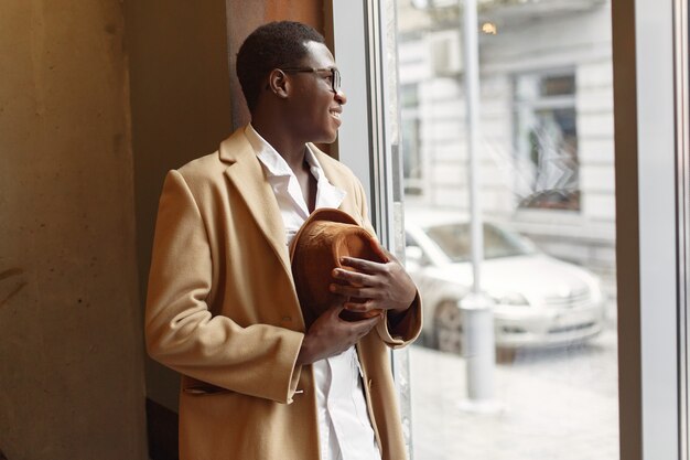 Hombre negro con un abrigo marrón de pie junto a la ventana