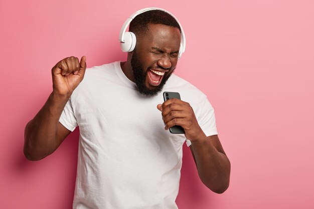El hombre negro sin afeitar, enérgico, canta con música, se mueve activamente, usa auriculares y una camiseta informal, posa sobre un fondo rosa, mantiene la boca abierta