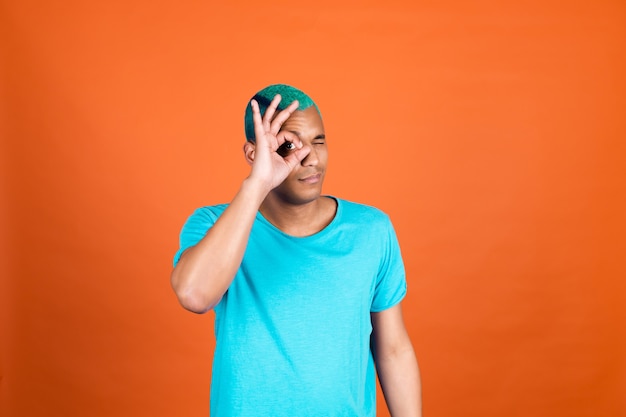 Hombre negro africano en casual en la pared naranja cabello azul que muestra un gesto de ok cubrir un ojo