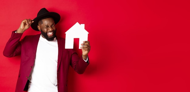 Hombre negro alegre de bienes raíces que muestra la casa de papel y el corredor recomendante sonriente de pie sobre rojo