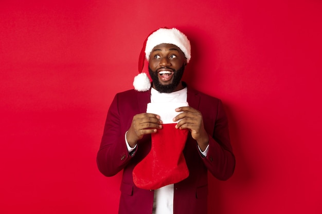 Hombre negro alegre mirando la esquina superior izquierda y sonriendo, sosteniendo el calcetín de Navidad con regalos, de pie sobre fondo rojo.