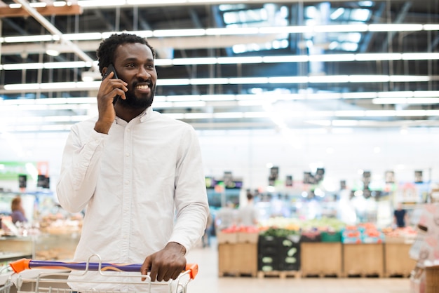 Hombre negro alegre que habla en el teléfono celular en la tienda de comestibles