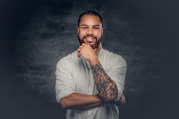 Hombre negro barbudo positivo con un tatuaje en los brazos cruzados, vestido con una camisa blanca.