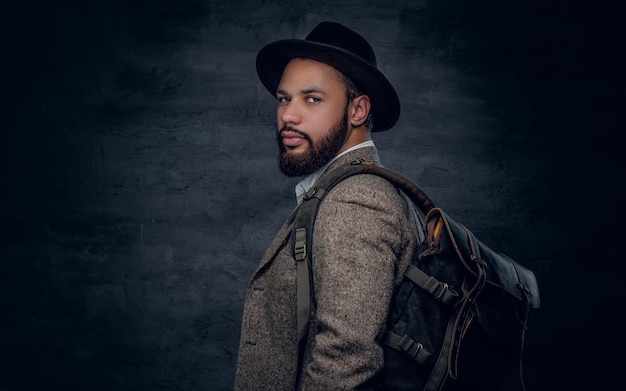 Foto gratuita hombre negro barbudo vestido con un traje de lana y un sombrero de fieltro sostiene una mochila.