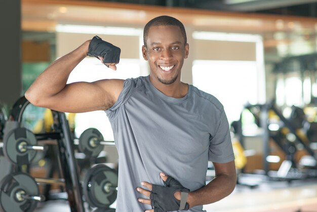 Hombre negro deportivo sonriente que muestra el bíceps en gimnasio