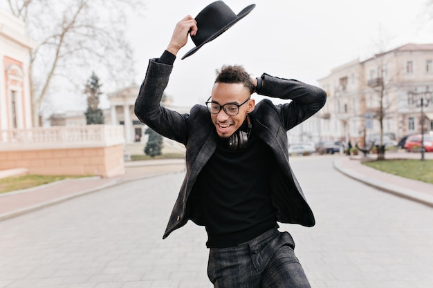 Hombre negro divertido jugando durante la sesión de fotos al aire libre en el parque. Chico africano escalofriante en traje elegante disfrutando de caminar por la calle en un día frío.