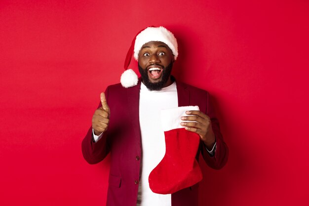 Hombre negro emocionado mostrando el pulgar hacia arriba en aprobación, sosteniendo un calcetín de Navidad con regalos navideños, sonriendo asombrado, de pie sobre fondo rojo.