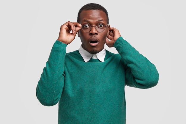 Hombre negro estupefacto con expresión facial asombrada, mantiene la mano en el borde de las gafas, vestido con un suéter verde informal