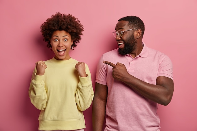 El hombre negro feliz con barba espesa señala a la mujer triunfante con los puños cerrados, celebra el éxito, exclama con alegría, expresa emociones positivas. Mirar