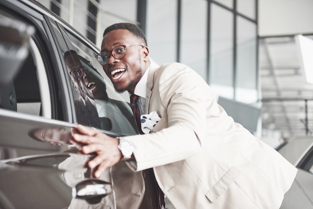 Hombre negro guapo en concesionario está abrazando a su nuevo coche y sonriendo.