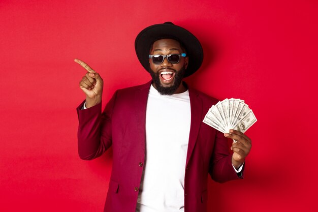 Hombre negro guapo y elegante señalando con el dedo a la izquierda mientras muestra dinero