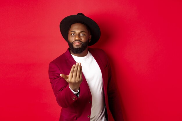 Hombre negro guapo y masculino pidiendo acercarse, atrayendo a dar un paso adelante, llamándote, de pie sobre fondo rojo