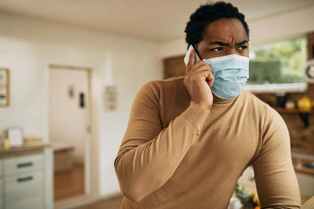 Hombre negro hablando por teléfono mientras usa una máscara protectora en casa