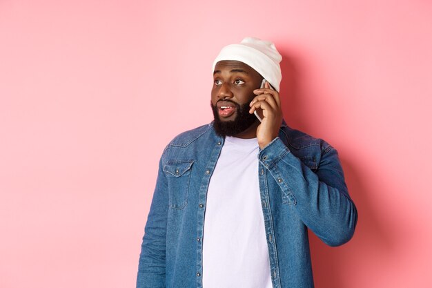 Hombre negro inconformista hablando por teléfono, mirando a la izquierda y teniendo una conversación móvil, de pie sobre fondo rosa