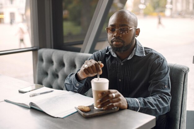 Hombre negro sentado en una cafetería y bebiendo un café con leche. Hombre con gafas de sol y camisa gris