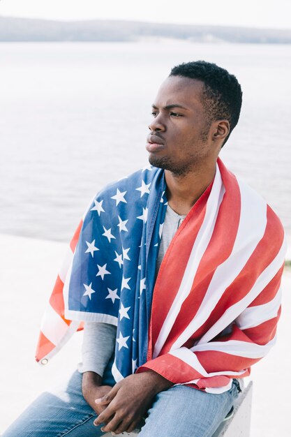Hombre negro serio envuelto en bandera estadounidense sentado en la playa