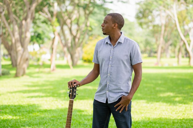 Hombre negro serio que sostiene la guitarra por el cabezal en el parque