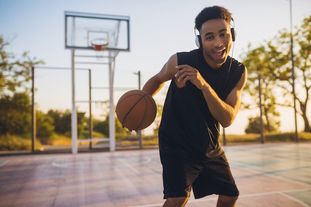 Hombre negro sonriente fresco haciendo deportes, jugando baloncesto al amanecer, escuchando música en auriculares, estilo de vida activo, mañana de verano