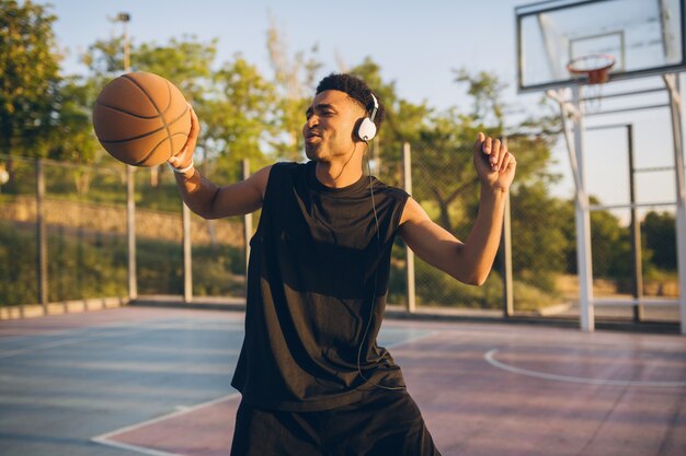 Hombre negro sonriente fresco haciendo deportes, jugando baloncesto al amanecer, escuchando música en auriculares, estilo de vida activo, mañana de verano