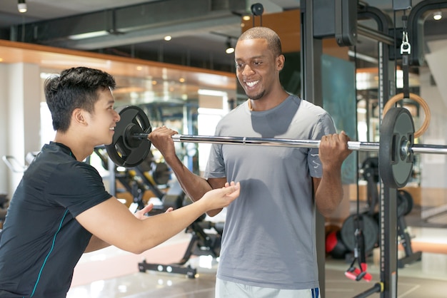 Hombre negro sonriente levantando barra con entrenador personal