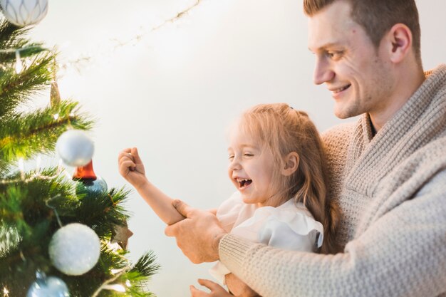 Hombre y niña riendo decorando árbol de navidad iluminado