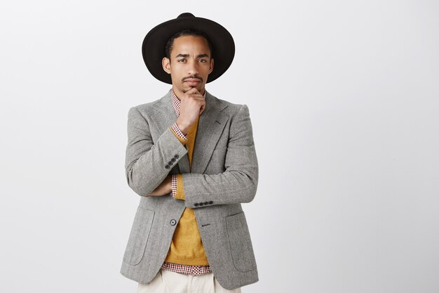 El hombre no está seguro de ti. Retrato de hombre guapo dudoso con chaqueta y sombrero de moda, sosteniendo la mano en la barbilla, frunciendo el ceño, mirando sospechoso mientras piensa, tiene dudas o incredulidad sobre la pared gris