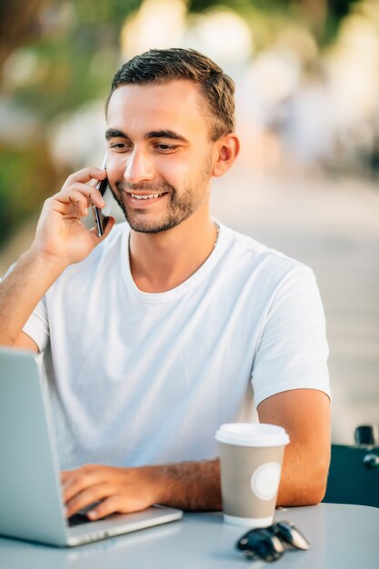 Hombre o estudiante inteligente sonriente exitoso joven en camisa casual, gafas sentado en la mesa, hablando por teléfono móvil en el parque de la ciudad usando laptop, trabajando al aire libre. Concepto de oficina móvil