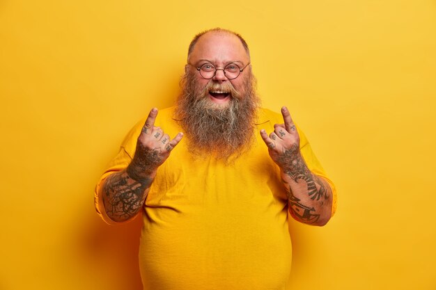 Hombre obeso gracioso con camiseta amarilla, muestra letrero de heavy metal, asiste al concierto de su banda de música favorita, tiene una gran barriga, brazos y barba tatuados, usa anteojos redondos. Gestos de fan de rock con sobrepeso en interiores