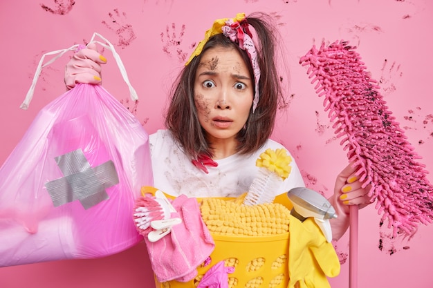 Hombre ocupado limpiando la casa recoge basura en una bolsa de polietileno sostiene poses de fregona cerca de la canasta de lavandería aislado en rosa