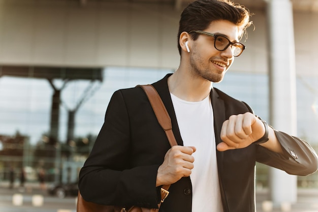 Foto gratuita hombre ocupado mira el reloj y sonríe retrato de un chico guapo moreno con chaqueta negra y camiseta blanca sosteniendo una mochila marrón