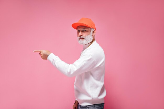 Hombre de ojos azules con gorra naranja que señala el lugar para el texto sobre fondo rosa Chico moderno con barba gris vestido con traje blanco posando