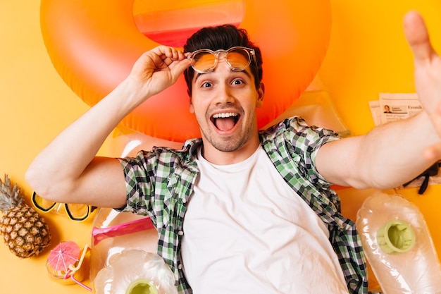 Hombre de ojos marrones con camiseta blanca y camisa a cuadros se quita las gafas de sol y se toma una selfie en un colchón inflable.