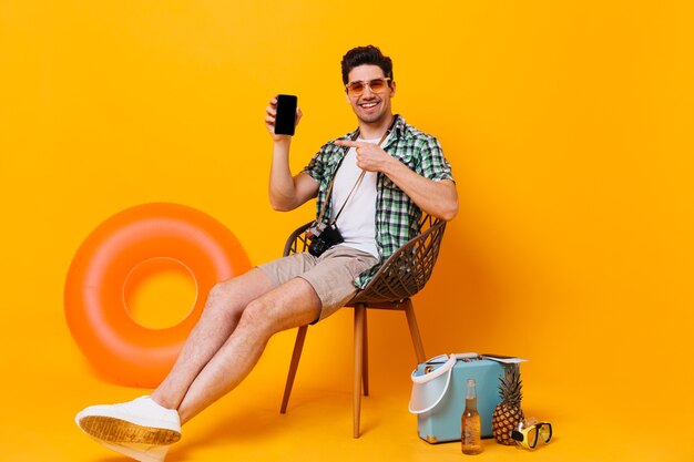 Hombre optimista en camisa verde y pantalón beige y mostrando en su teléfono inteligente. Retrato de chico con gafas de sol sentado en una silla con maleta, cerveza, círculo inflable