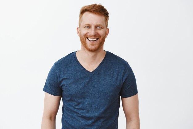 Hombre ordinario guapo alegre con cabello pelirrojo y cerdas en camiseta azul, sonriendo ampliamente