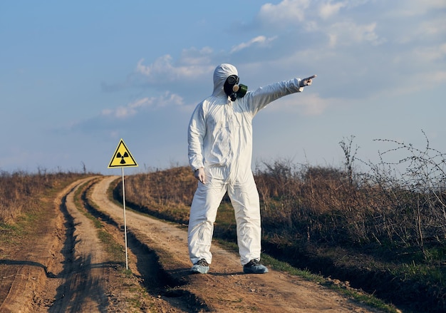 Hombre en overol protector y máscara de gas de pie en la carretera del pueblo en el campo junto a la señal de precaución