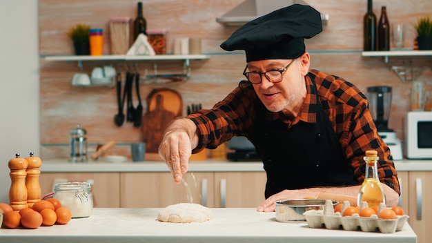 Hombre de panadería tamizando la harina sobre la masa en la mesa en la cocina de casa. chef anciano jubilado con bonete y uniforme rociando, tamizando, esparciendo ingredientes rew con pan y pizza casera para hornear a mano.