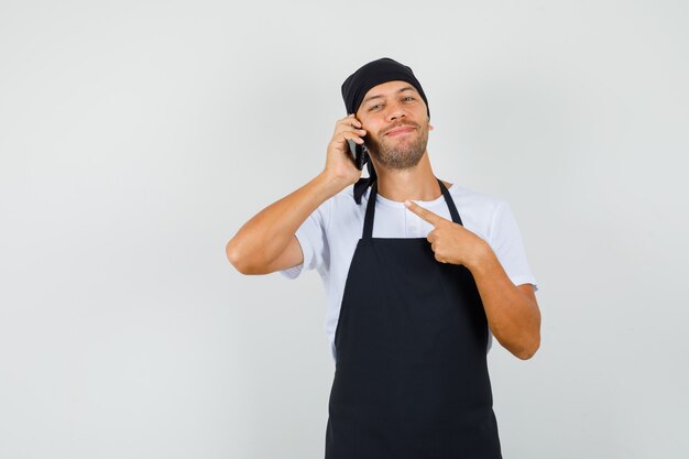 Hombre panadero apuntando al teléfono móvil en camiseta, delantal y mirando alegre.