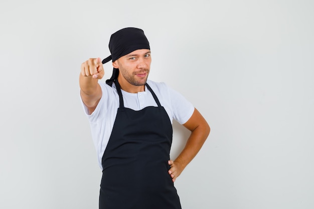 Hombre panadero apuntando a la cámara en camiseta, delantal y mirando confiado
