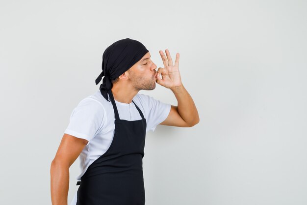 Hombre panadero en camiseta, delantal mostrando gesto delicioso besando los dedos y mirando encantado