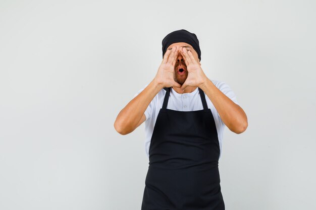 Hombre panadero en camiseta, delantal susurrando chismes con las manos cerca de la boca abierta