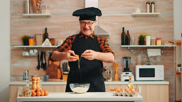 Hombre panadero experimentado craking huevos para hornear vistiendo delantal disfrutando de la afición. Chef anciano jubilado con bonete mezclando a mano, amasando en un tazón de vidrio ingredientes de pastelería preparando tarta casera