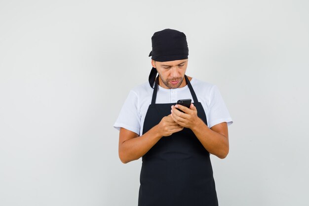 Hombre panadero con teléfono celular en camiseta, delantal y mirando ocupado