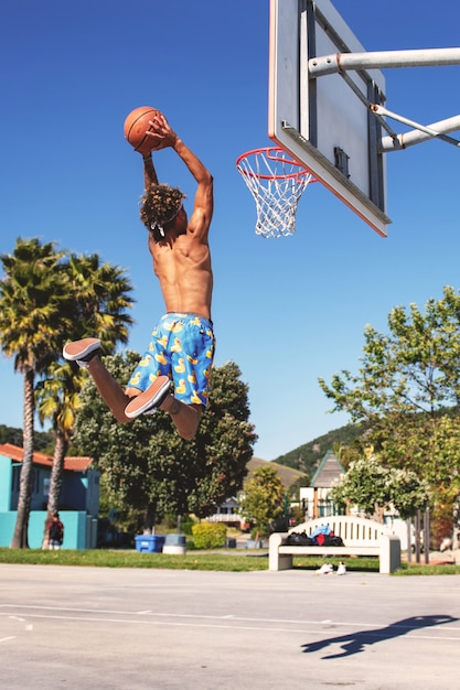 Hombre con pantalones cortos azules y amarillos haciendo un mate en la cancha de baloncesto durante el día