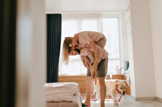 Un hombre con pantalones cortos de color caqui sostiene a su joven esposa boca arriba mientras su perro camina junto a él. Los amantes disfrutan de un fin de semana sin preocupaciones en su apartamento.