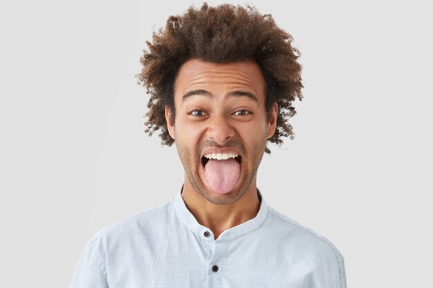 Hombre con peinado afro muestra la lengua mientras nota algo repugnante, hace muecas, demuestra un carácter obstinado