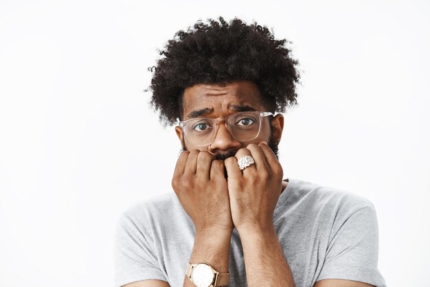 Hombre con peinado afro en reloj y gafas mordiéndose las uñas levantando las cejas tonto y tímido, teniendo miedo de tener susto en la pared gris.