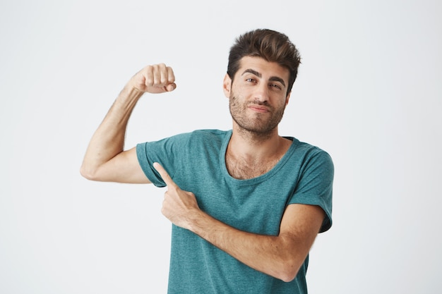 Hombre con peinado elegante y barba frunciendo el ceño, demostrando lo fuerte que es. Apto chico apuntando con su dedo a sus bíceps. Macho man mostrándose